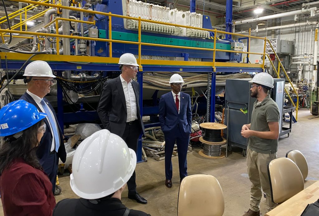 men and women wearing hard hats stand in front of industrial equipment answering questions