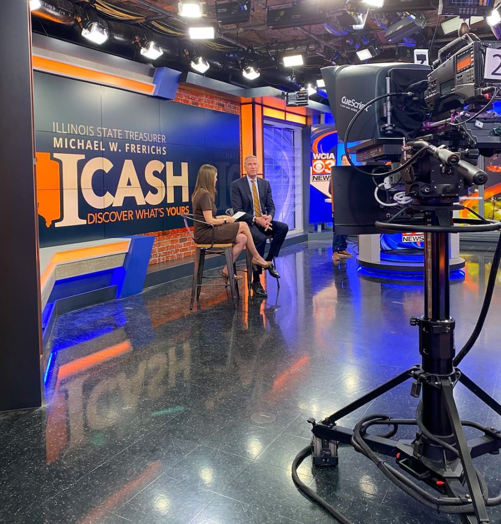 a television camera in the foreground with a man and a woman on stools in the background having a conversation