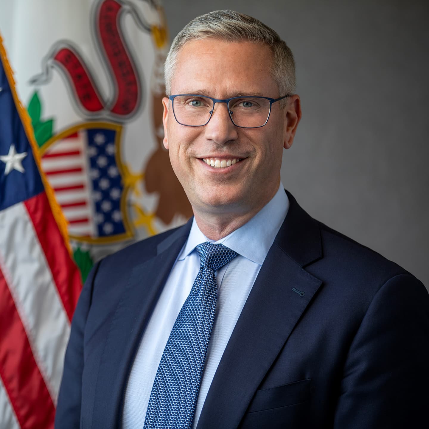 Portrait of an elected official wearing a blue suit, smiling, with the Illinois flag behind them.