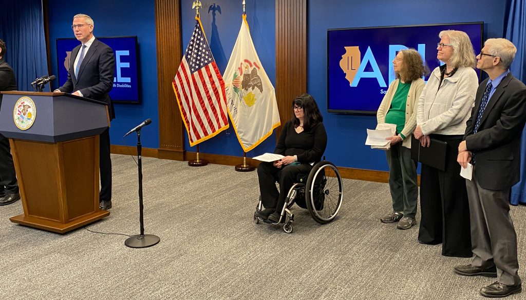 Treasurer Frerichs stands at a podium as four people, including one using a wheelchair, are beside him
