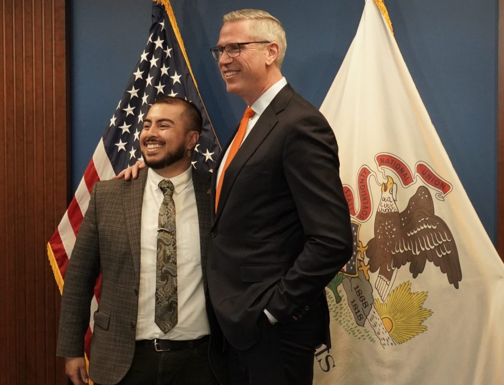 two men in suits standing in front of the illinois flag and united states flag