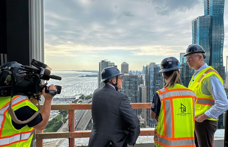 Treasurer Frerichs and 2 other people standing outside at a construction site as the Chicago skyline is in the background, and a TV camera person is on the left