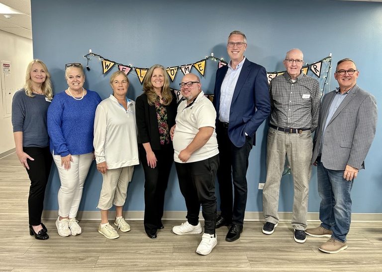 Eight people pose in front of a blue wall at Synapse House. Treasurer Frerichs is third from right.
