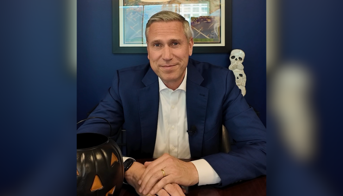 Treasurer Michael Frerichs smiling at the camera while seated at a desk with an orange and black decorative pumpkin. Above his left shoulder are two small skull decorations.
